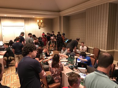 A few of the 2017 voting village. There are about 30 people crowding a fairly small room. There are voting machines and other computer parts strewn about the room and many people are just standing around.