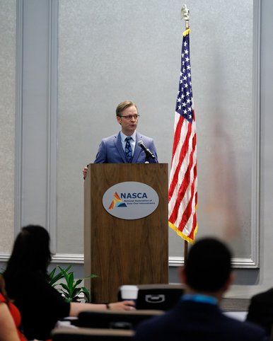 A man in a sky-blue three-piece suit with a light blue shirt and a dark blue tie, speaking from behind a podium at a conference