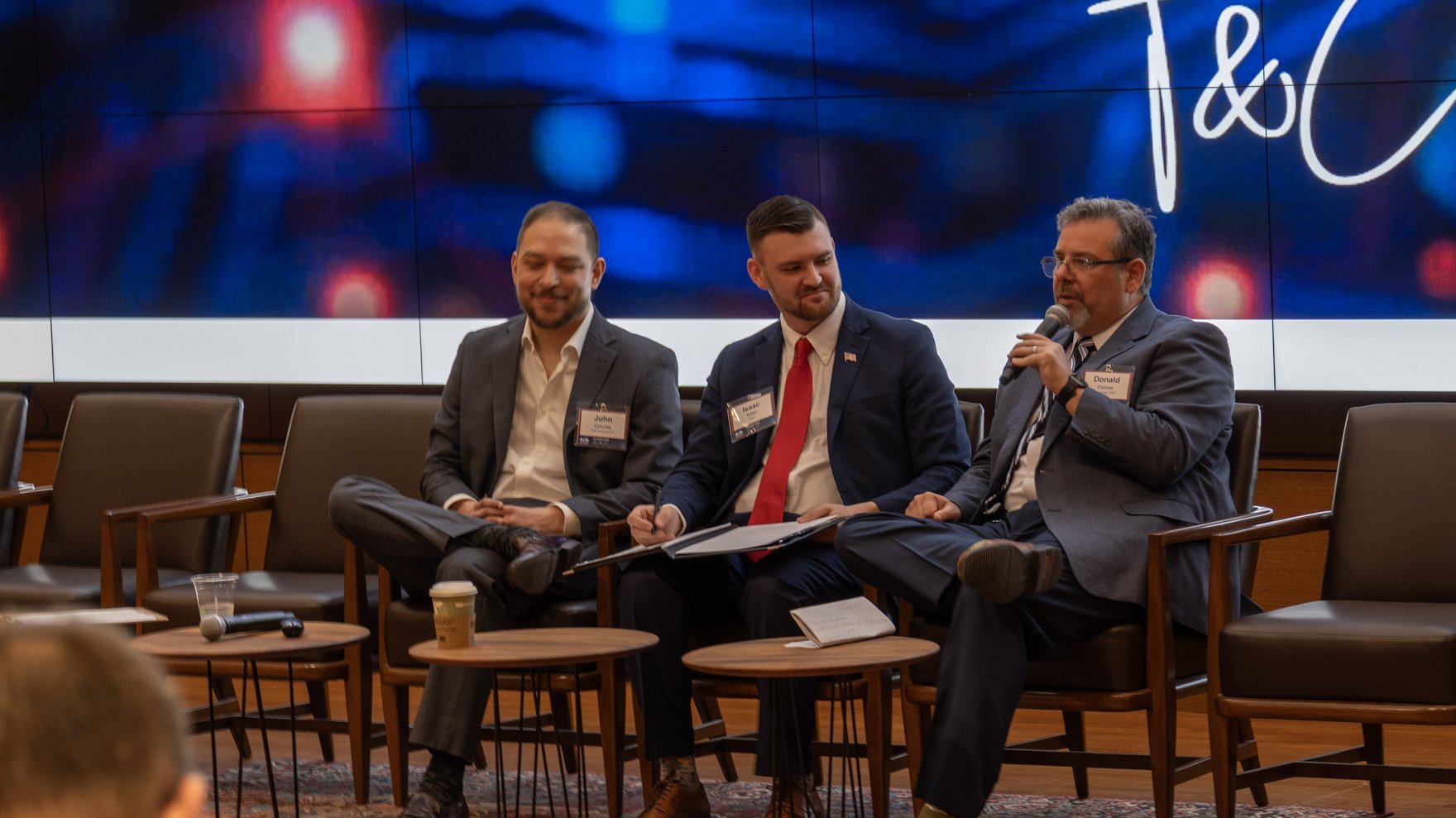 Three people in suits are seated on a panel on a stage in front of a large screen.