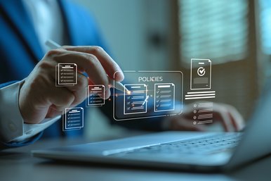 A cropped image of a person in front of a laptop, showing only the person's hands. There are line icons representing policy documents floating over the keyboard. The person is interacting with the icons with a stylus.